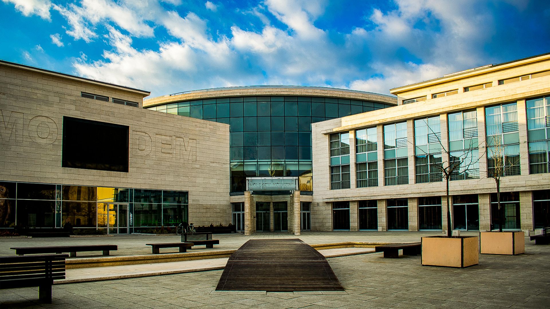 The modern glass and stone facade of the MODEM Center for Modern and Contemporary Art in Debrecen, Hungary, under a partly cloudy sky.