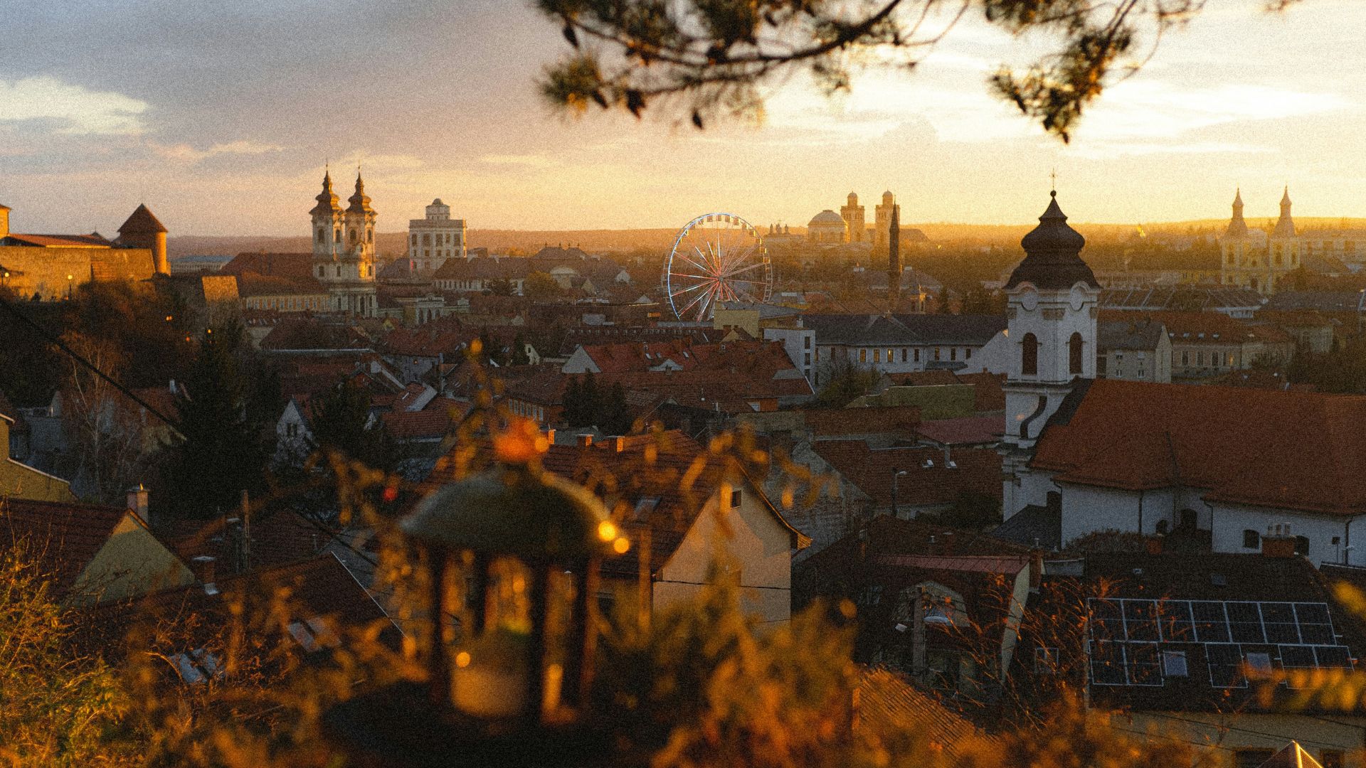 The historic cityscape of Eger, Hungary, at sunset, with church towers, a Ferris wheel, and rooftops glowing in warm evening light.