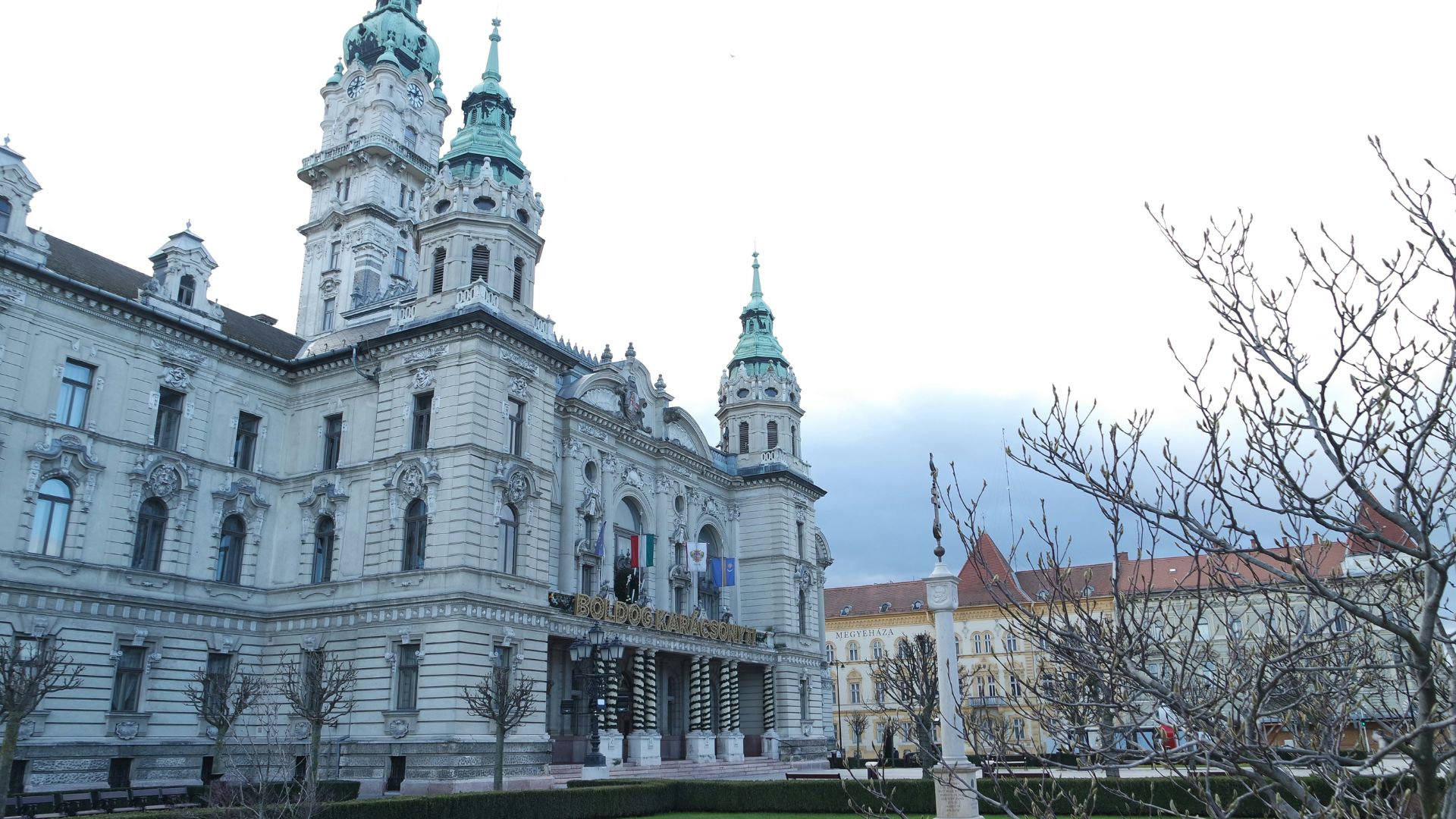 The ornate facade of the Gyor town hall, featuring twin green domes, with bare trees and historic buildings in the surrounding square.