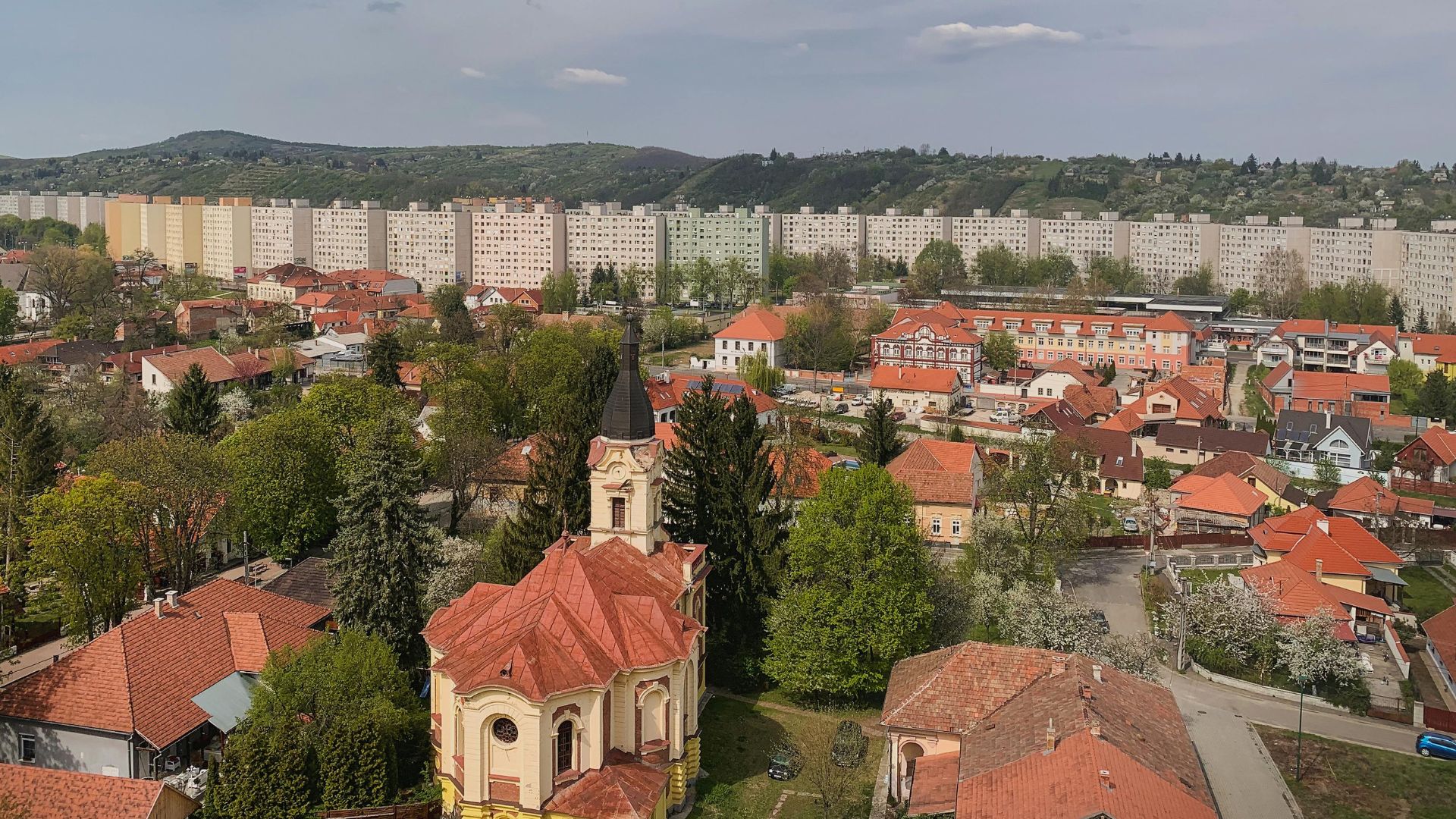 An aerial view of Miskolc, Hungary, showing a mix of historic homes and newer apartment buildings, with green hills in the background.