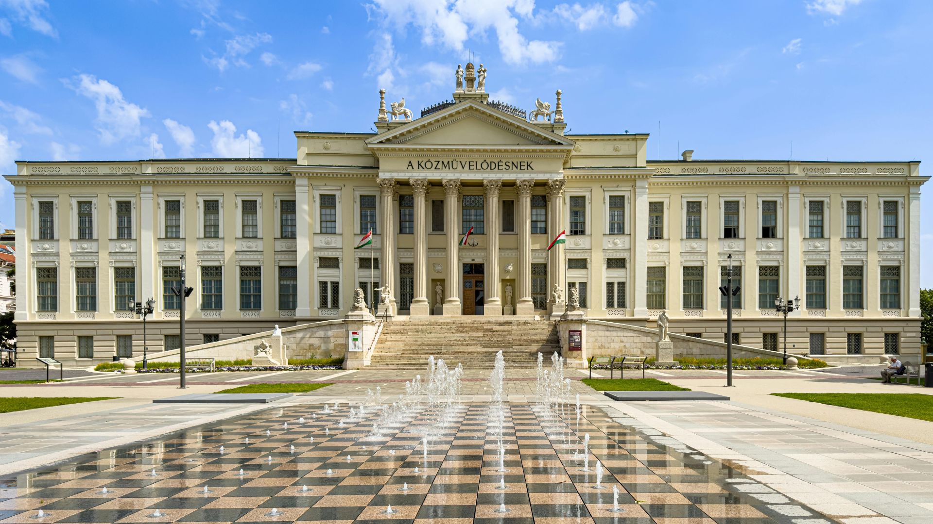 The grand facade of the Móra Ferenc Museum in Szeged, Hungary, with a symmetrical water fountain in the foreground and blue skies above.