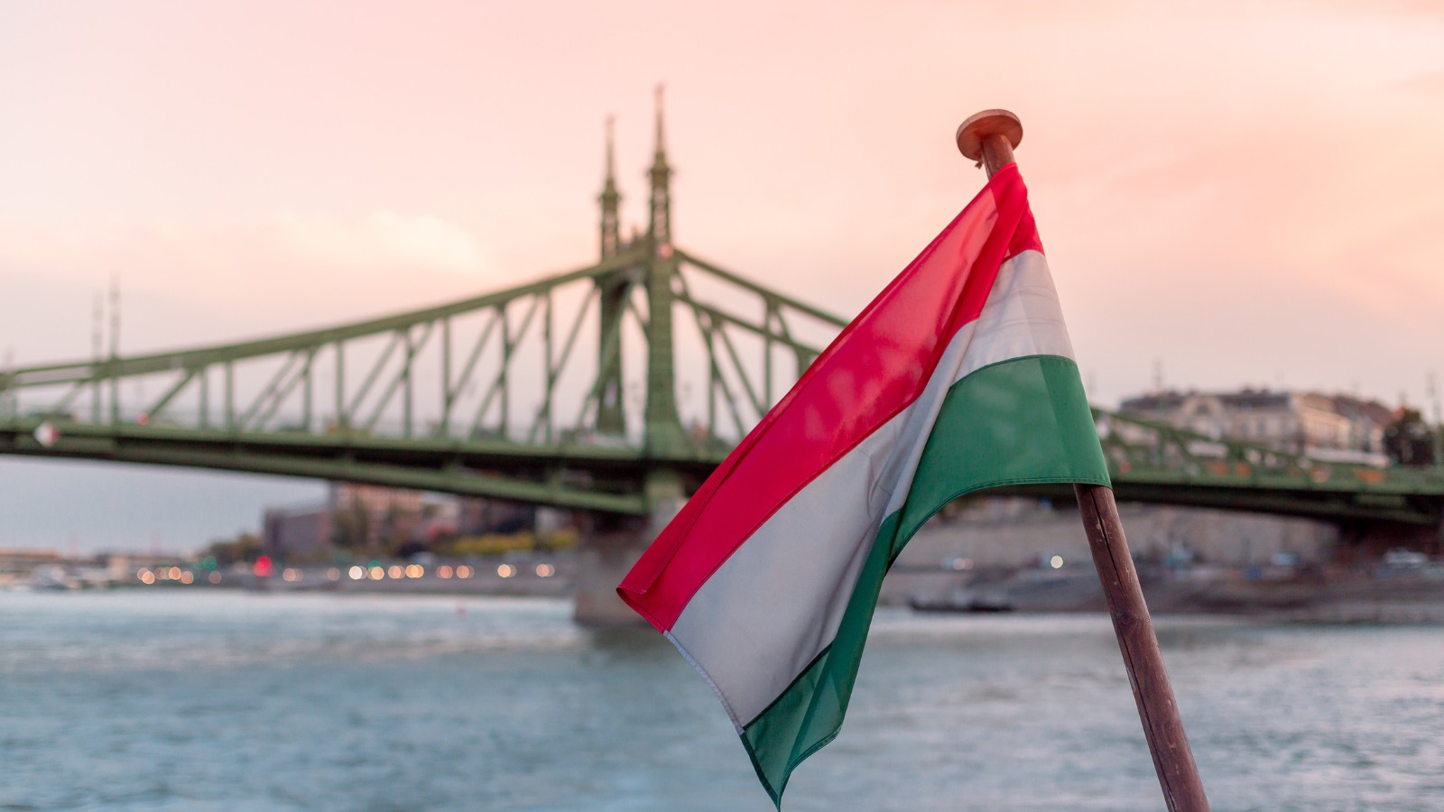 Hungarian flag waving in the foreground with Liberty Bridge (Szabadság híd) and the Danube River in the background at sunset in Budapest.